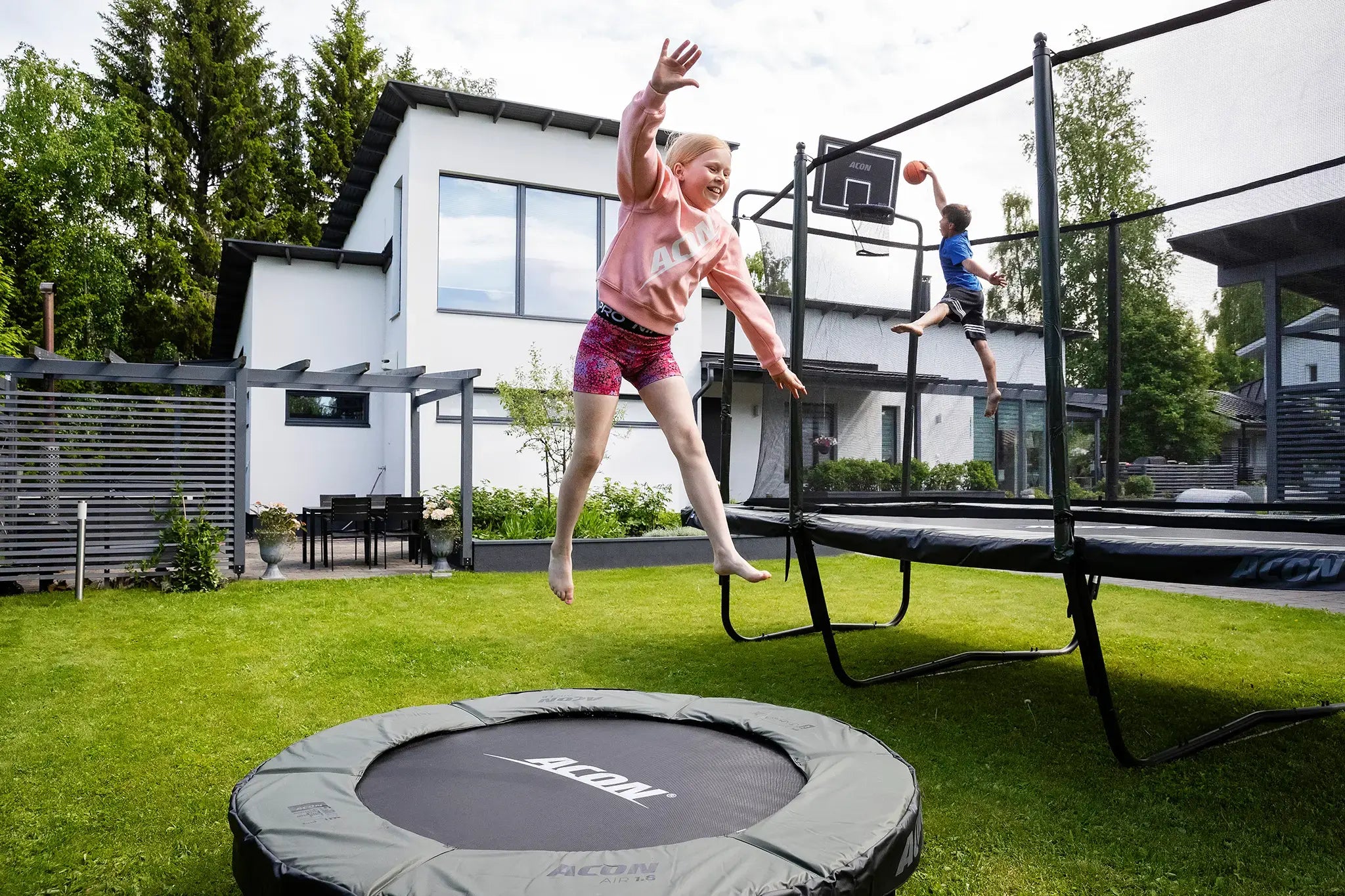 Suuren talon takapihalla kaksi Acon trampoliinia ja lapsia hyppimässä. Edustalla pienempi pihatrampoliini, ja taustalla suuri suorakulmainen trampoliini turvaverkolla.